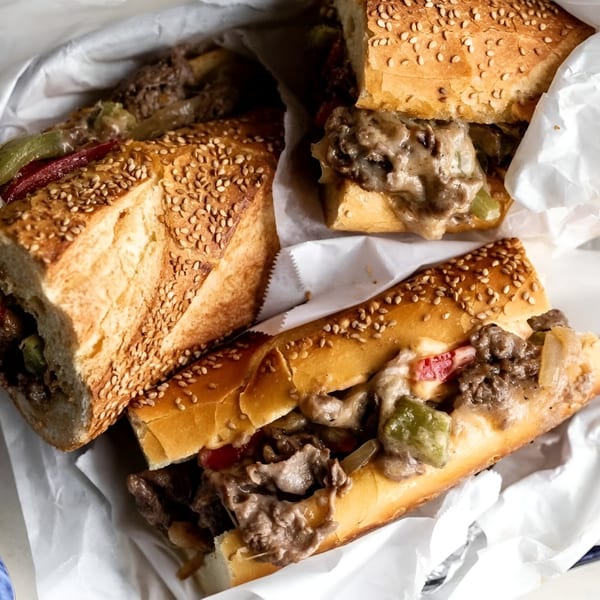 Three cheesesteak sandwiches with meat, cheese, peppers, and onions on sesame seed rolls in white paper.