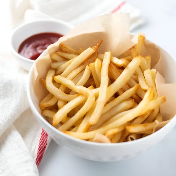 A white bowl filled with golden french fries and a side of ketchup on a white surface.